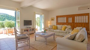 Ein geräumiges Wohnzimmer mit einer großen Couch, einem Couchtisch und einem Fernseher. Ein Holzschrank und ein Essbereich sind durch eine offene Tür sichtbar.