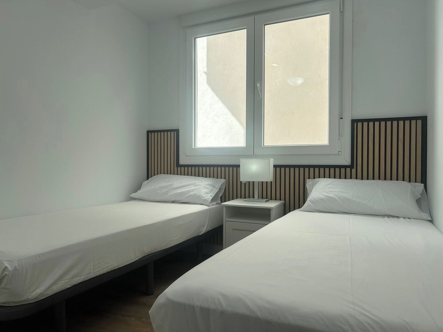 A twin room with two single beds, a nightstand with a lamp, and a window with blinds.