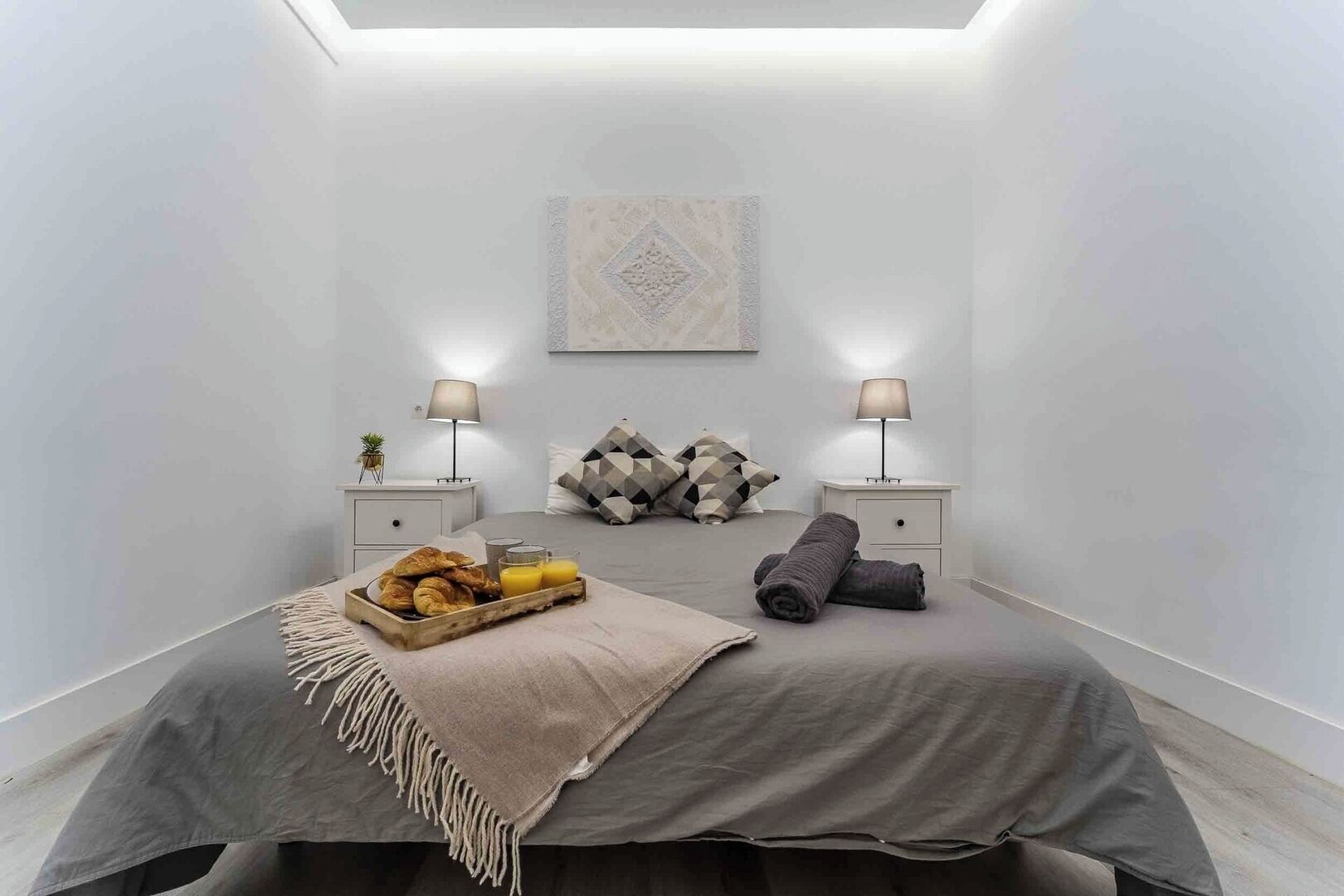 Un dormitorio moderno con tragaluz, una cama con cubrecama gris, dos mesitas de noche con lámparas, una bandeja de croissants y una manta enrollada.