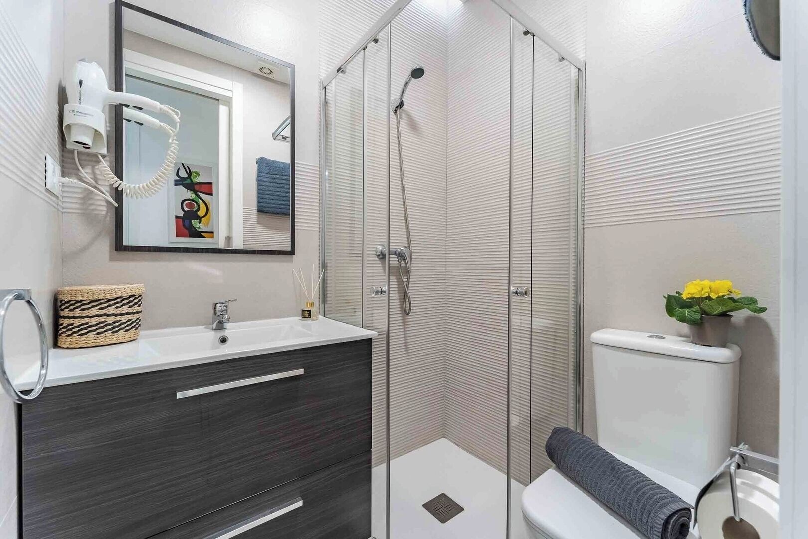 Un baño moderno con cabina de ducha de vidrio, un lavamanos blanco sobre un mueble oscuro y un inodoro con una toalla y flores.