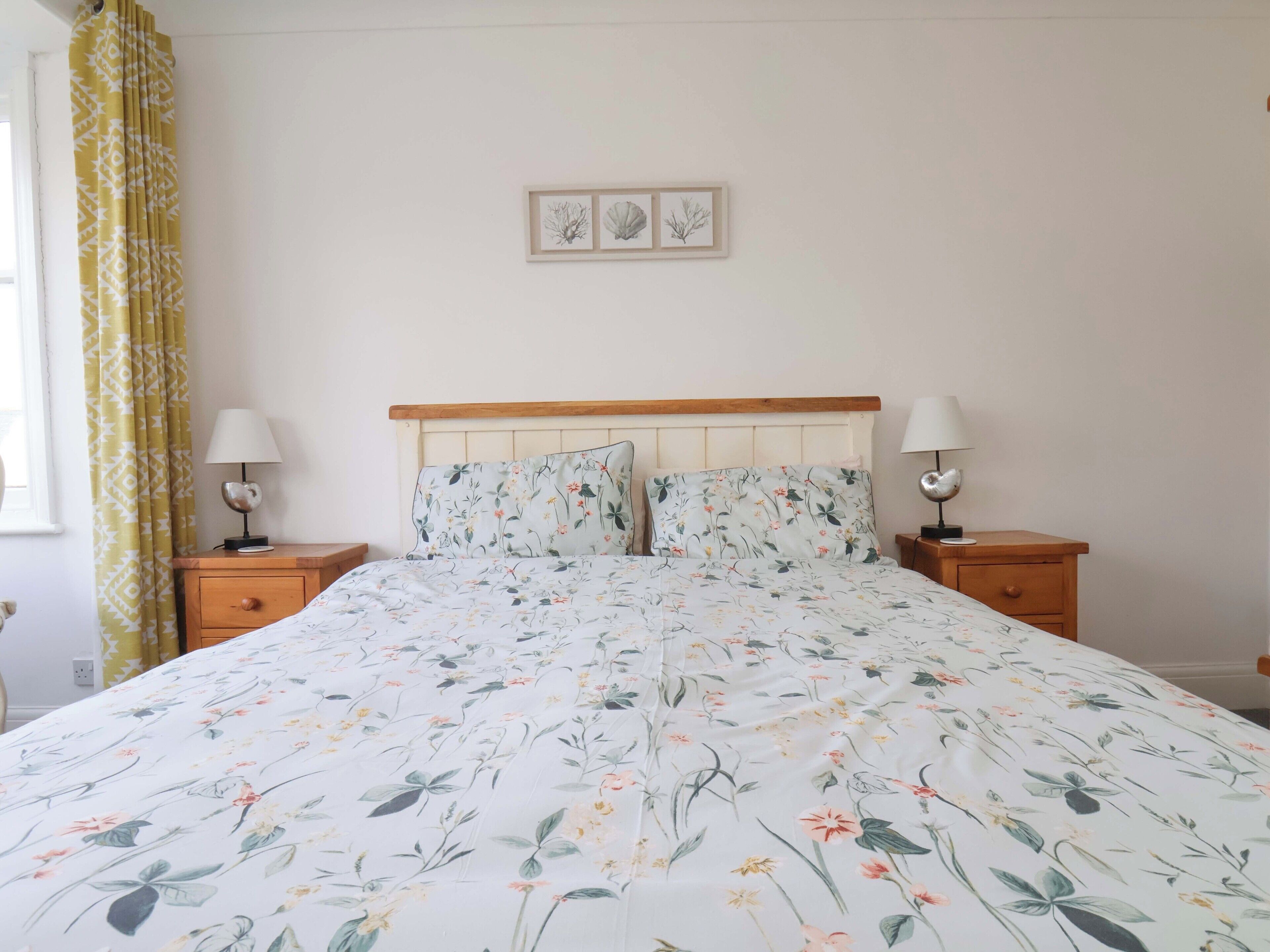 Quarto com colcha florida, cabeceira de madeira, duas mesas de cabeceira com luminárias e uma janela com cortinas.