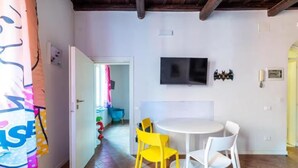 A room with a white round table, yellow and white chairs, a flat-screen TV on the wall, and a wooden ceiling. - Welcome in Trastevere apartment (Roma)