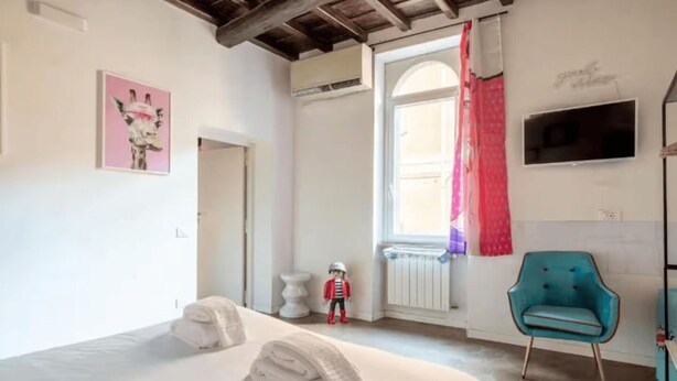 A bedroom with a bed, a blue chair, a small table, a radiator, and a window with curtains. - Welcome in Trastevere apartment (Roma)