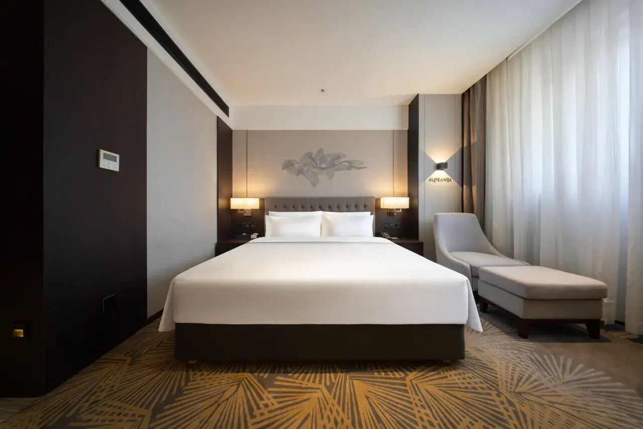 A hotel room with a large bed, two bedside lamps, a chair, and a large window with curtains.