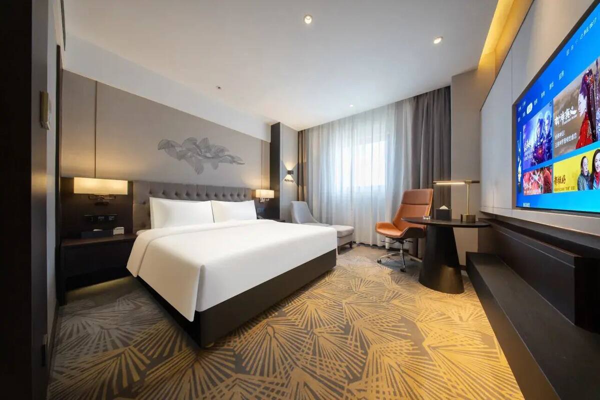 Ein Hotelzimmer mit einem großen Bett, einem Schreibtisch, einem Sessel sowie einem Fernseher und einem an der Wand befestigten Bildschirm, der Inhalte anzeigt.