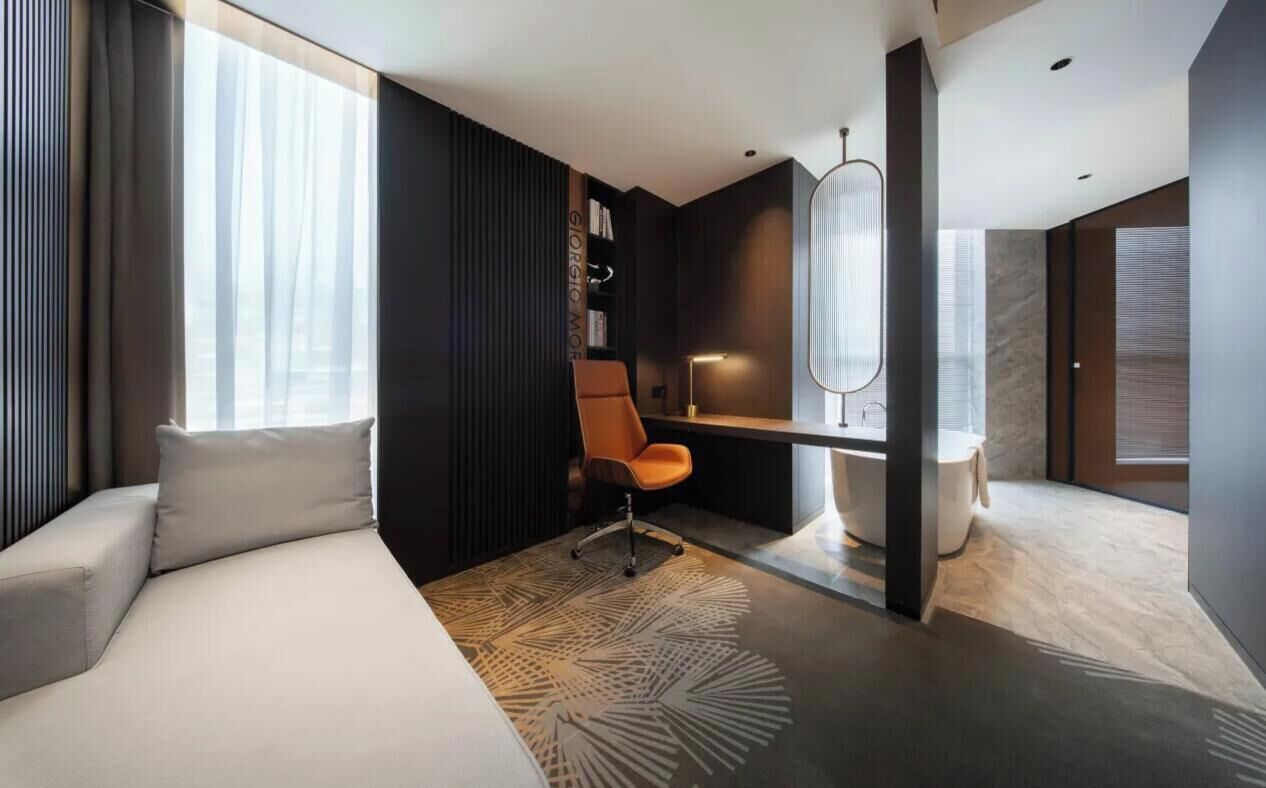 A modern hotel room with a bed, a desk, a chair, a small table, and a large window.