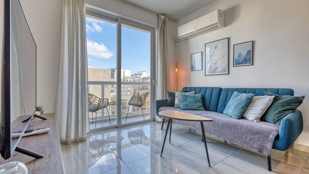 A modern living room with a blue sofa, a coffee table, and a large sliding glass door leading to a balcony. - Sofia`s Designer Apartment (St. Julian`s)