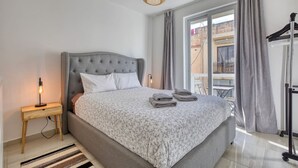A bedroom with a grey upholstered bed, a bedside table with a lamp, a window with curtains, and a view of the outside balcony. - Sofia`s Designer Apartment (St. Julian`s)
