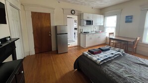 A compact room with a bed, a kitchenette, a dining table, and a door leading to another room. - Bright Studio Near Boston — Transit Access — Washer/Dryer (Malden)