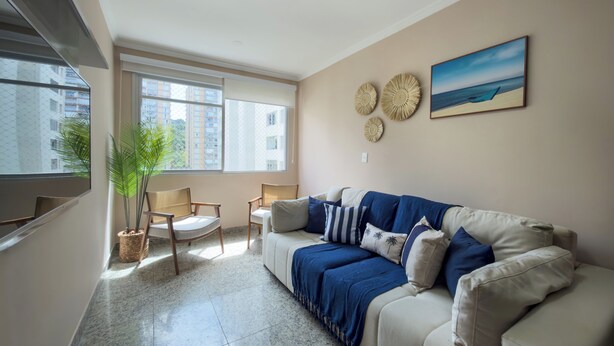 A living room with a beige sofa, a blue throw blanket, and a painting of a beach scene. There are two chairs and a potted plant. - Your refuge in Guarujá, just steps from the beach! BRA (Guarujá)