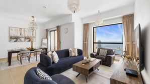 Ein modernes Wohnzimmer mit einer Couch, Sesseln, einem Couchtisch und einem Fernseher. Es gibt einen Essbereich mit Tisch und Stühlen, und ein großes Fenster mit Blick auf das Meer.