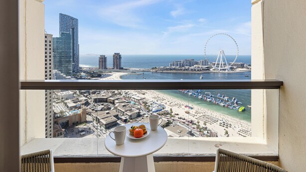 Ein Balkon mit Tisch, Blick auf einen Strand, ein Riesenrad und hohe Gebäude.