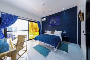Un dormitorio moderno con una cama grande, un ventilador de techo, vista a una habitación amarilla a través de una puerta de vidrio y una alfombra con diseño azul y blanco.