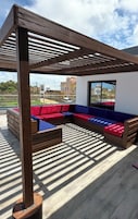 Un patio en la azotea con una pérgola de madera, una zona de estar con cojines rojos y azules, y vistas de la ciudad.