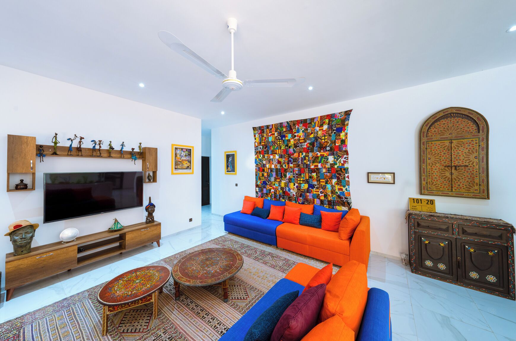 A modern living room with a flat-screen TV, a blue and orange sofa, a patterned rug, and a ceiling fan.