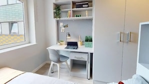 A bedroom with a bed, a desk with a chair, a wardrobe, and a window with blinds. - Student Only Highbury (London)