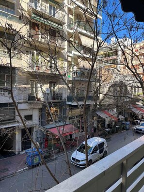 Um edifício residencial de vários andares com varandas, uma rua com carros estacionados e uma loja pequena com toldo vermelho.