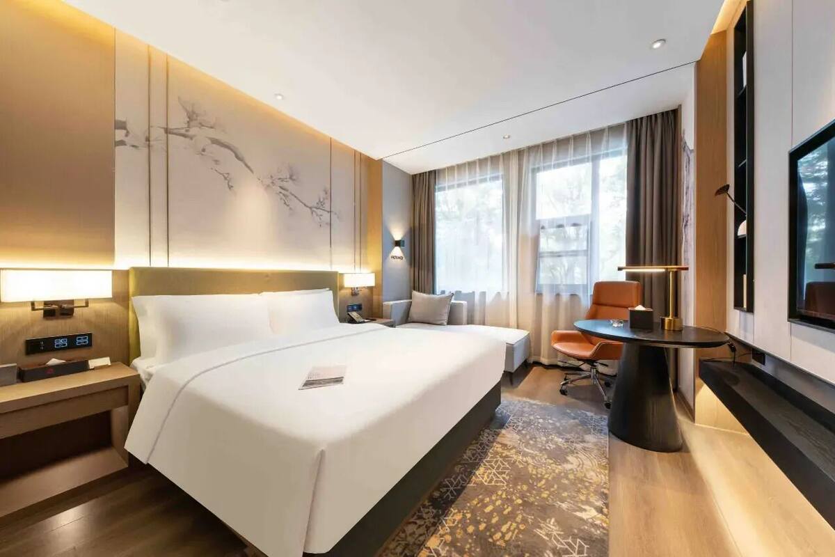 A hotel room with a large bed, bedside tables, a desk, a chair, a TV, and a window with curtains.