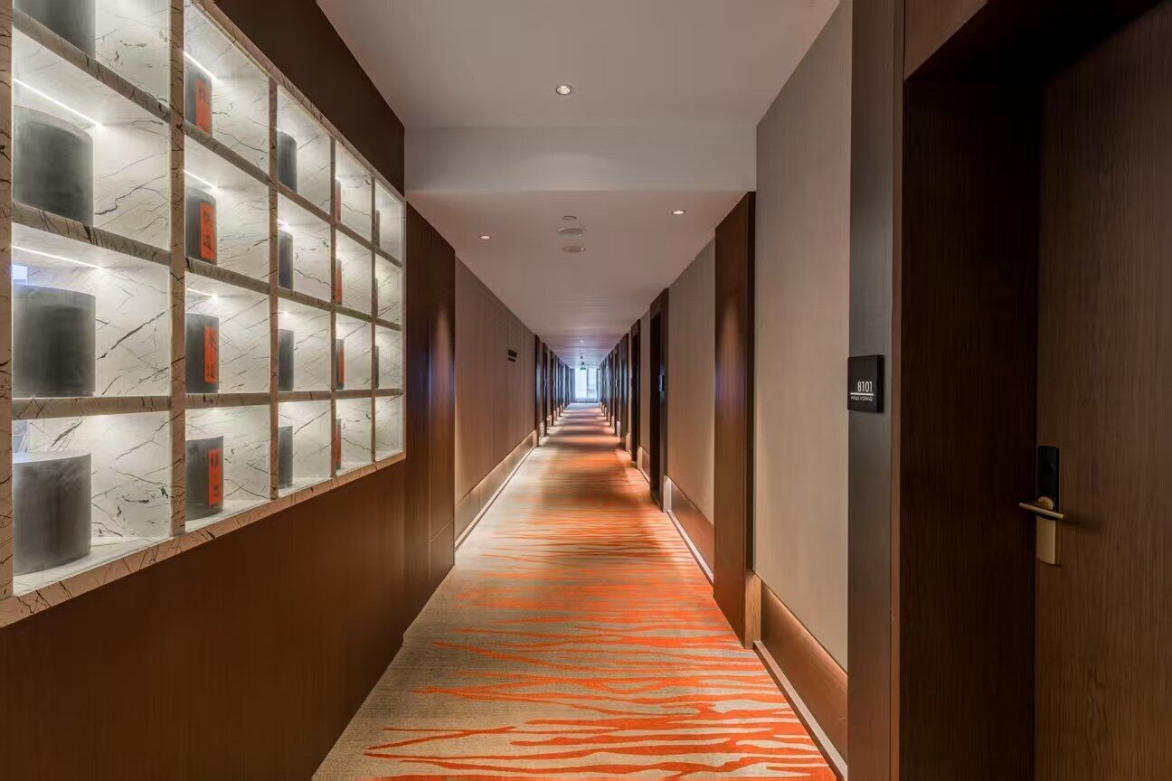 Un pasillo de hotel con alfombra estampada, paredes de madera y un separador de vidrio con paneles iluminados.