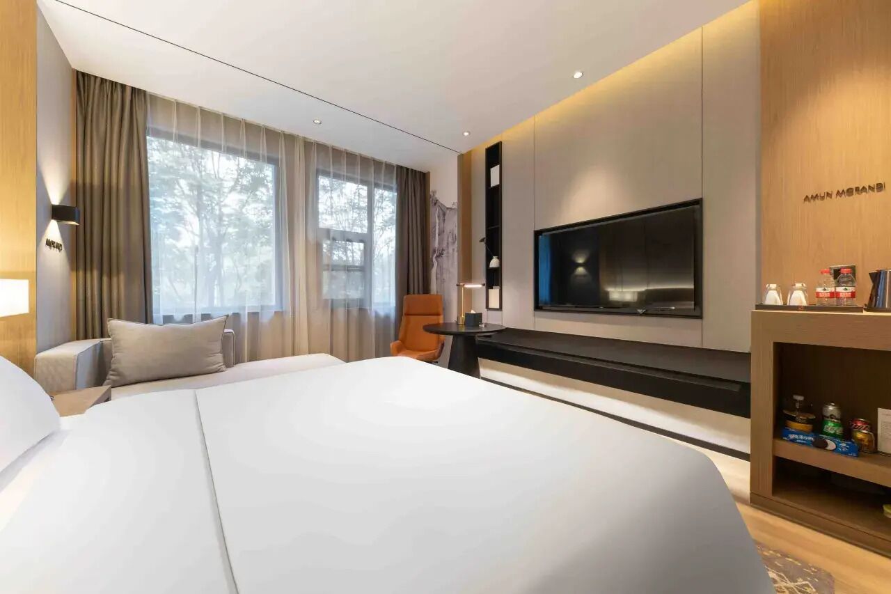 Ett modernt hotellrum med en stor säng, en platt-TV som är monterad på väggen, en sittgrupp med en stol och ett litet bord samt ett stort fönster med gardiner.
