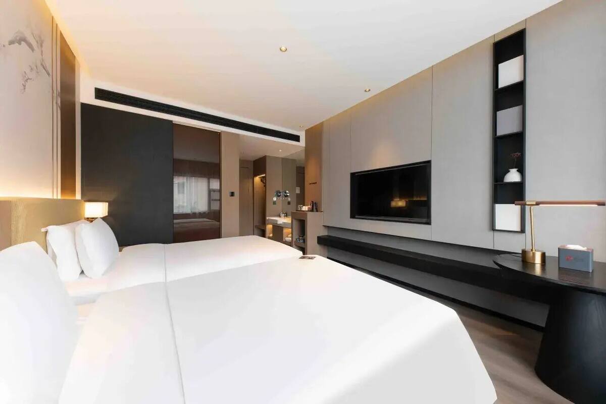 A modern hotel room with a large bed, a flat-screen TV mounted on the wall, a bedside table, and a built-in shelf.