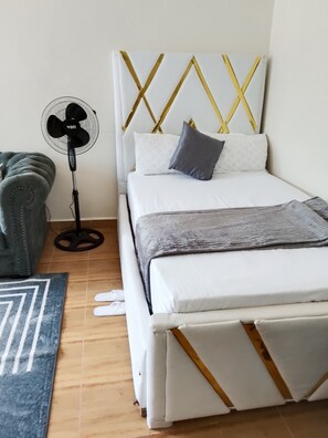 Un dormitorio ordenado con cama, un ventilador de pie, un sofá gris y una alfombra estampada.