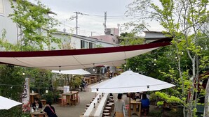 Um café ao ar livre com mesas e cadeiras, cercado por vegetação e edifícios.