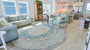 Un salon spacieux avec un canapé, des fauteuils et une table basse. On y trouve un grand tapis, une bibliothèque et un coin repas avec une table et des chaises. La cuisine est visible au fond, avec des appareils modernes et un lustre.