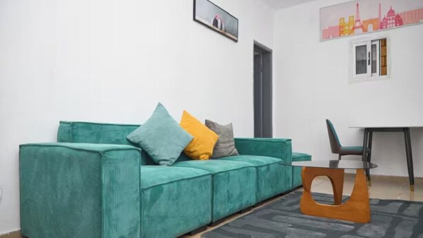 Un divano color petrolio con cuscini, un tavolino in legno e un tappeto grigio in una stanza dalle pareti bianche e un dipinto con vista sulla città.