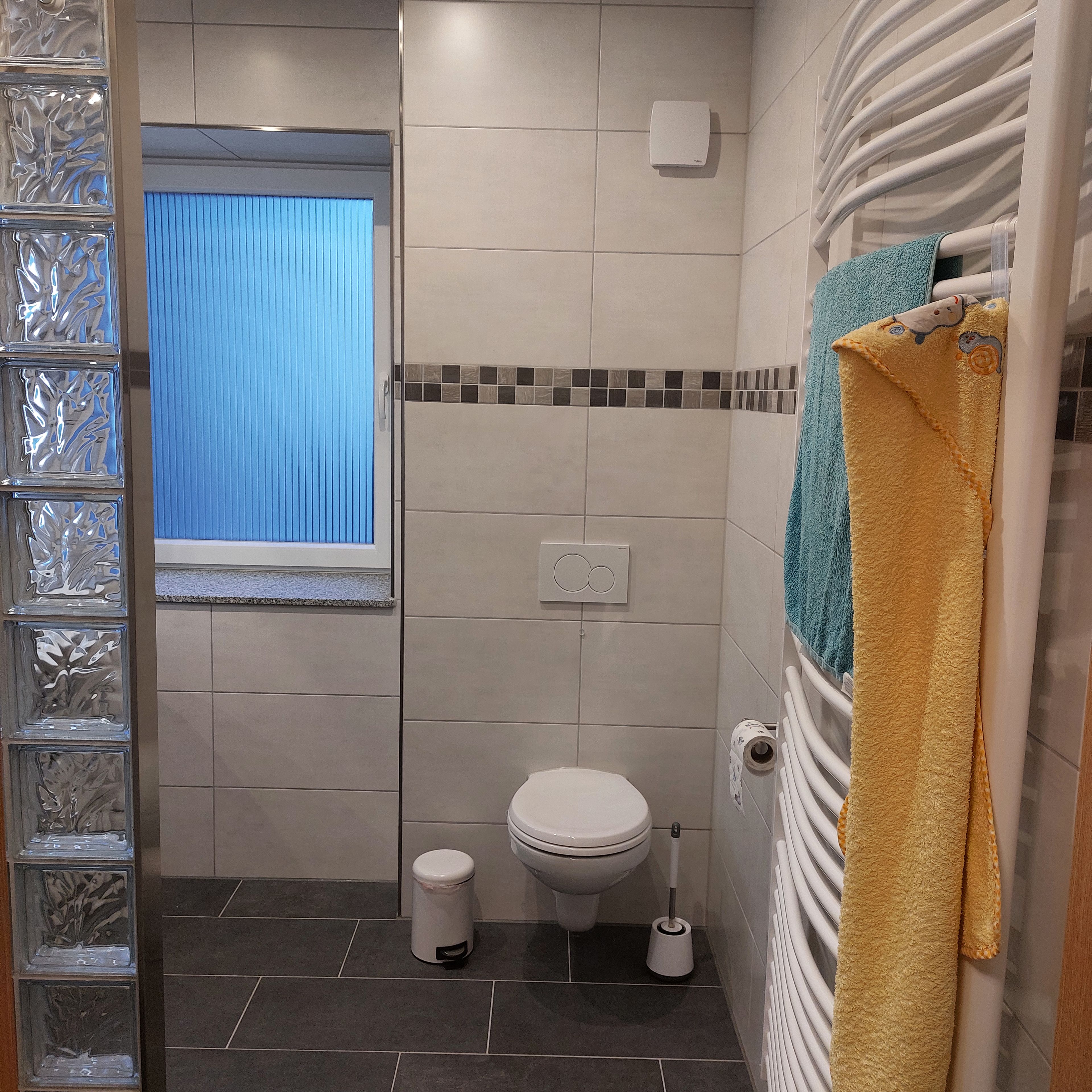 Un baño con inodoro, bidé, cubo de basura, toallero y toalla, y una ventana con persianas.
