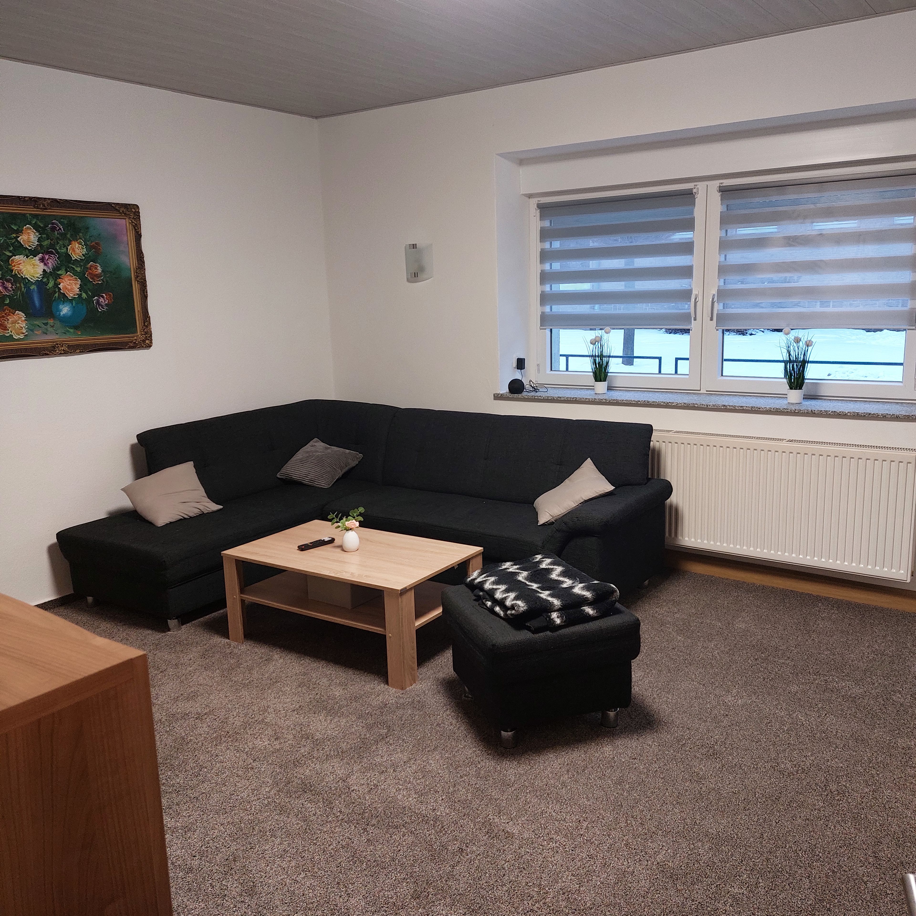 Una sala de estar con un sofá negro, una mesa de centro de madera y un reposapiés negro. Hay un cuadro en la pared y una ventana con persianas.