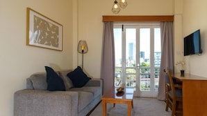 En stue med en grå sofa, et træ sofabord, et fjernsyn på væggen og en balkon med udsigt over byen.
