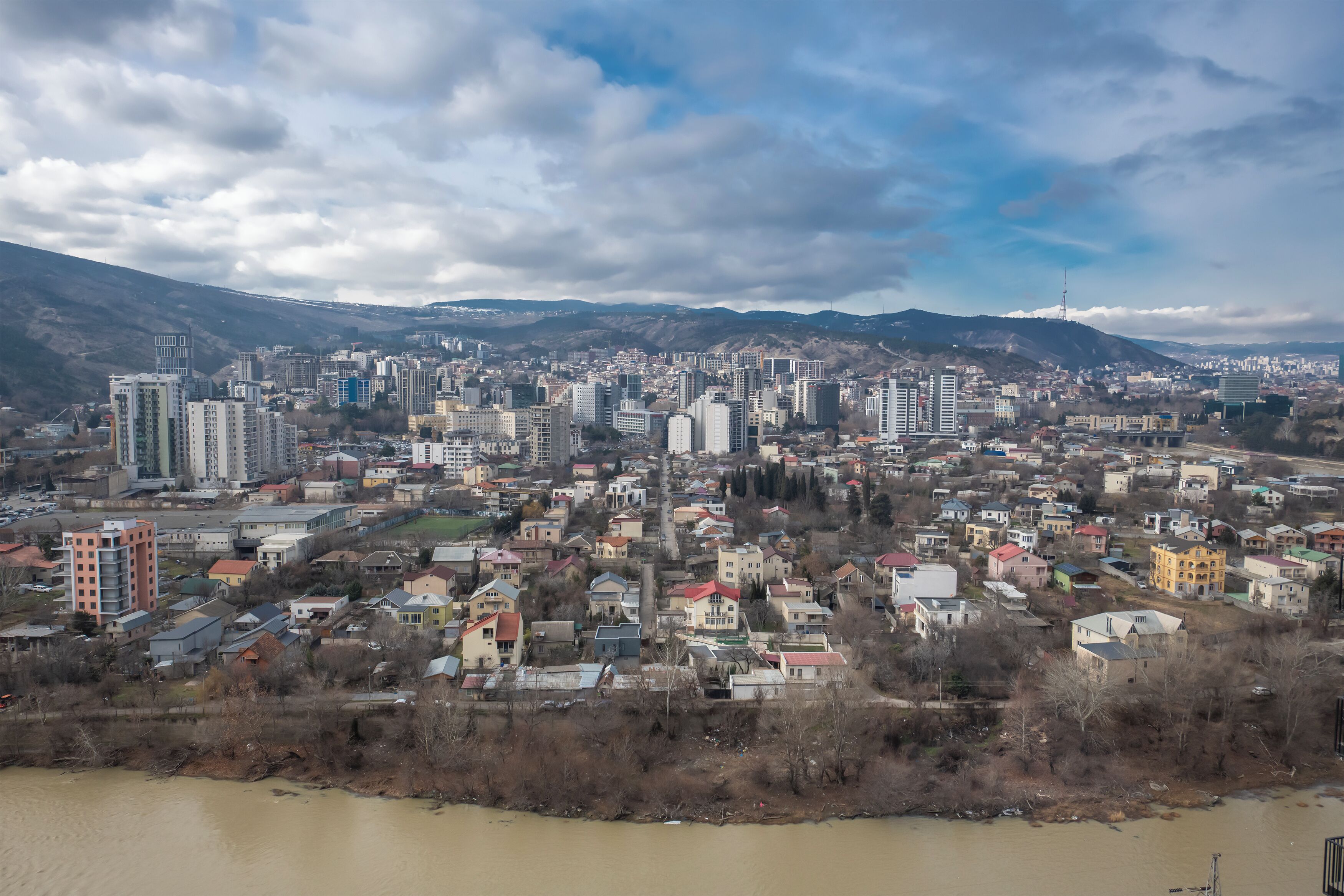 A cityscape with a river in the foreground, surrounded by residential and commercial buildings, and a mountain range in the background.