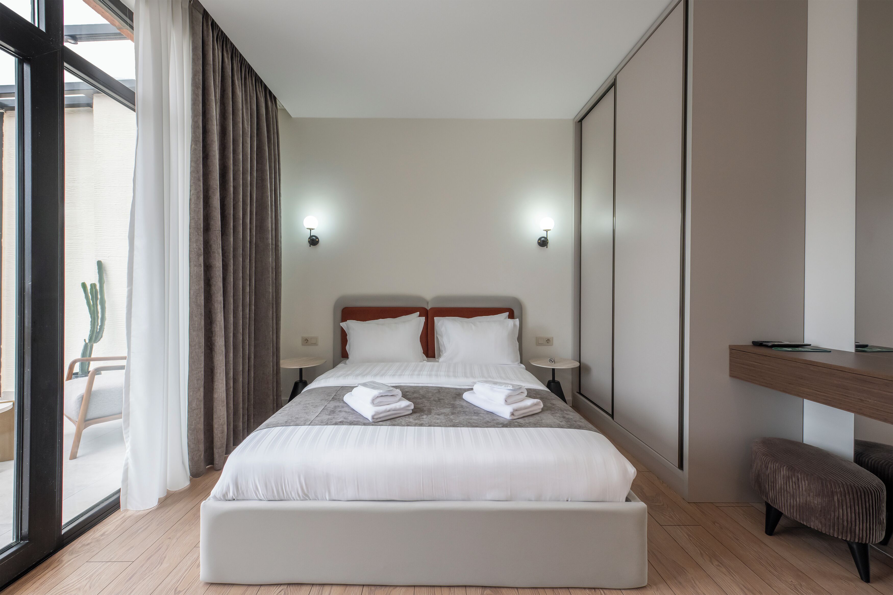Une chambre d’hôtel moderne avec un grand lit, deux tables de chevet, un petit banc et une porte coulissante donnant sur un balcon.