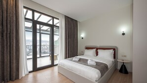 Une chambre d’hôtel avec un grand lit, une table ronde et une vue sur le paysage urbain, visible à travers une porte vitrée.