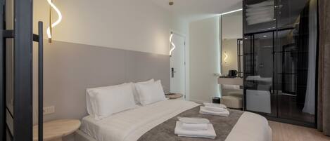 A modern hotel room with a large bed, a bedside table, a built-in shower, and a glass-enclosed walk-in closet.