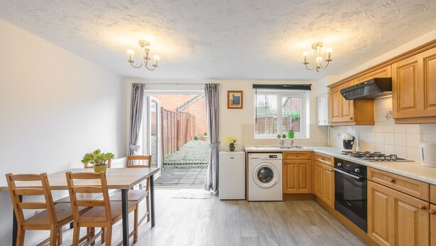 A compact kitchen with wooden cabinets, a built-in oven, a washing machine, and a dining area with a table and chairs. - Host and Stay - The Rowton (Norwich)
