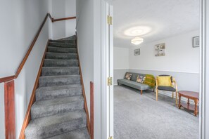A staircase with carpeted steps and wooden handrails, leading to a living area with a sofa, chairs, and a small table. - Host and Stay - The Rowton (Norwich)