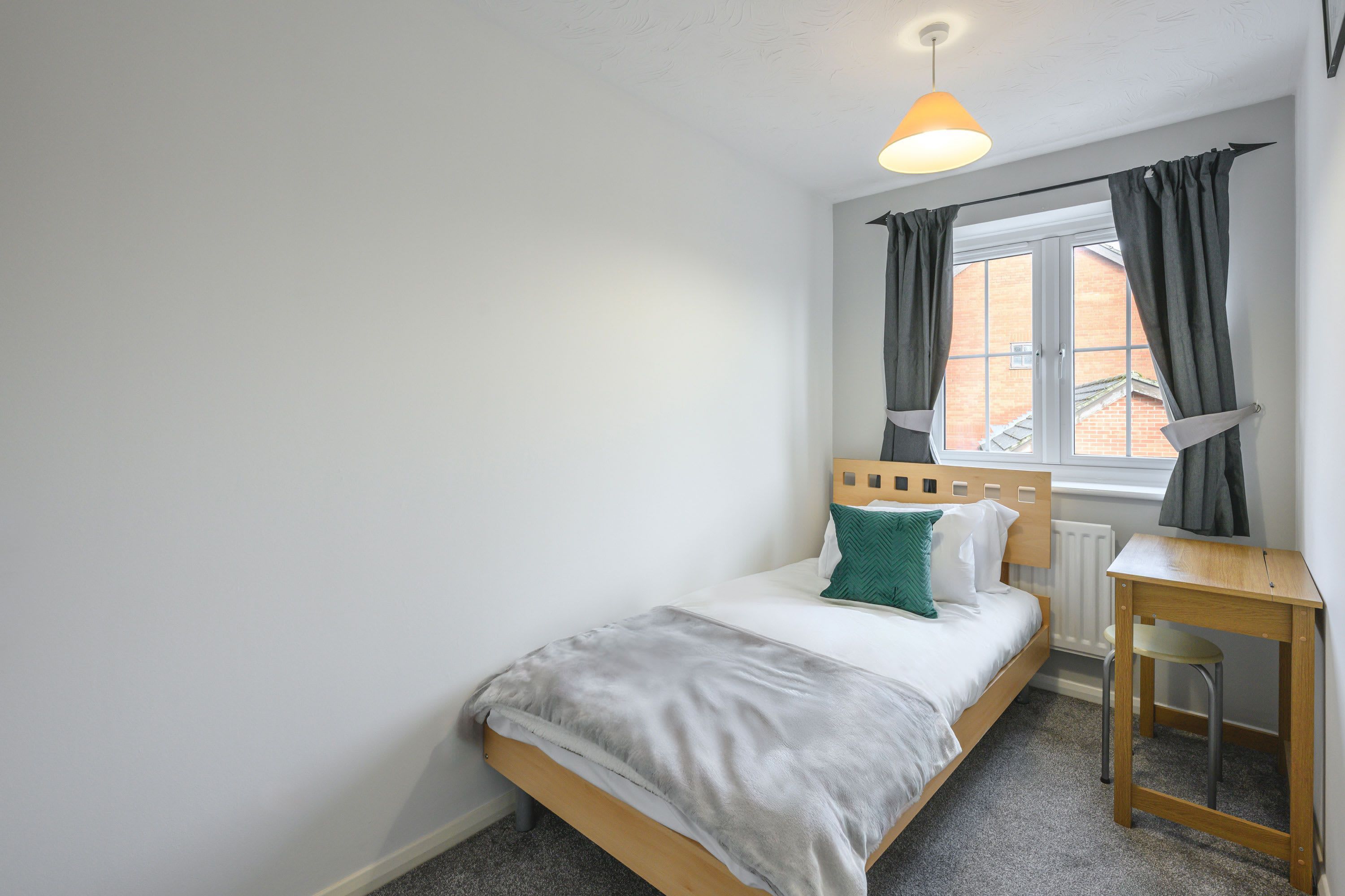 Une chambre avec un lit simple, un bureau en bois, une chaise, une fenêtre avec des rideaux et un luminaire suspendu.