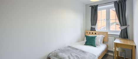 A bedroom with a single bed, a wooden desk, a chair, a window with curtains, and a hanging light fixture.