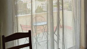 Ein Zimmer mit großem Fenster, das mit leichtem Stoff drapiert ist, ein Holzsessel und ein kleiner Tisch.
