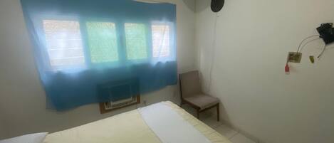 Ein Zimmer mit einem Bett, einem Sessel, einem Wandventilator und einem Fenster mit blauem Vorhang.