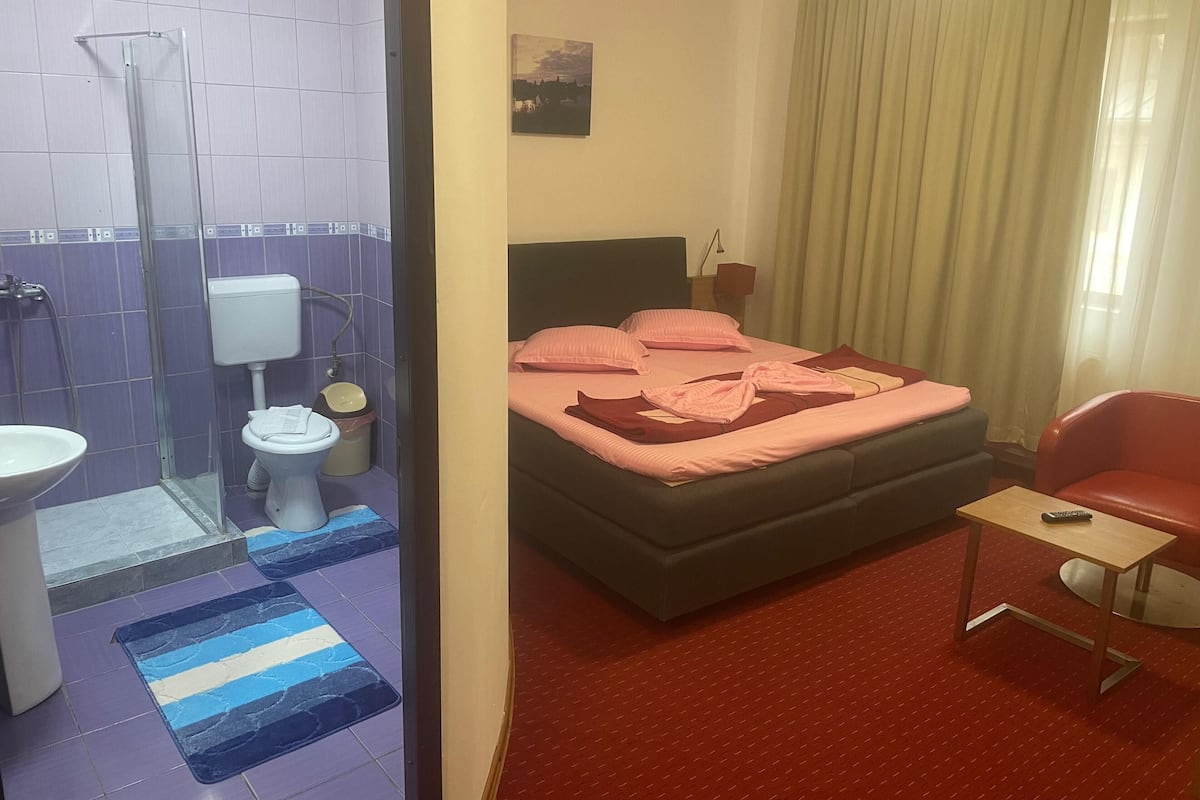 A hotel room with a bed, a small table, a red chair, a bathroom with a shower, toilet, and sink.
