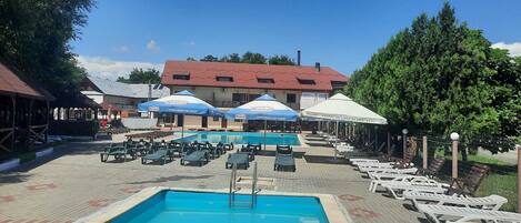 A pool area with lounge chairs and umbrellas, a building with a red roof, and a fenced garden area.