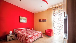 Un dormitorio con colcha de cama estampada en rojo y blanco, una silla roja, una mesita de noche con lámpara y un cuadro en la pared.