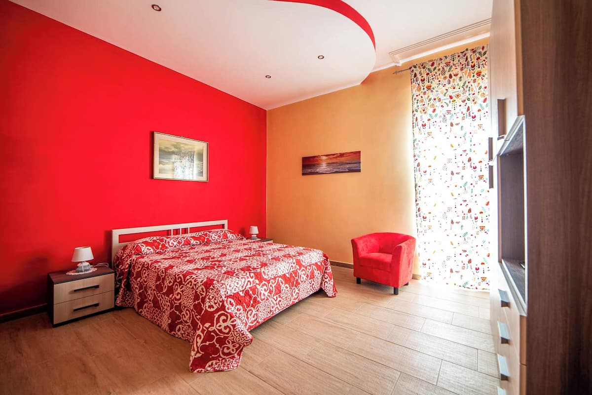 A bedroom with a red and white patterned bedspread, a red chair, a nightstand with a lamp, and a painting on the wall.