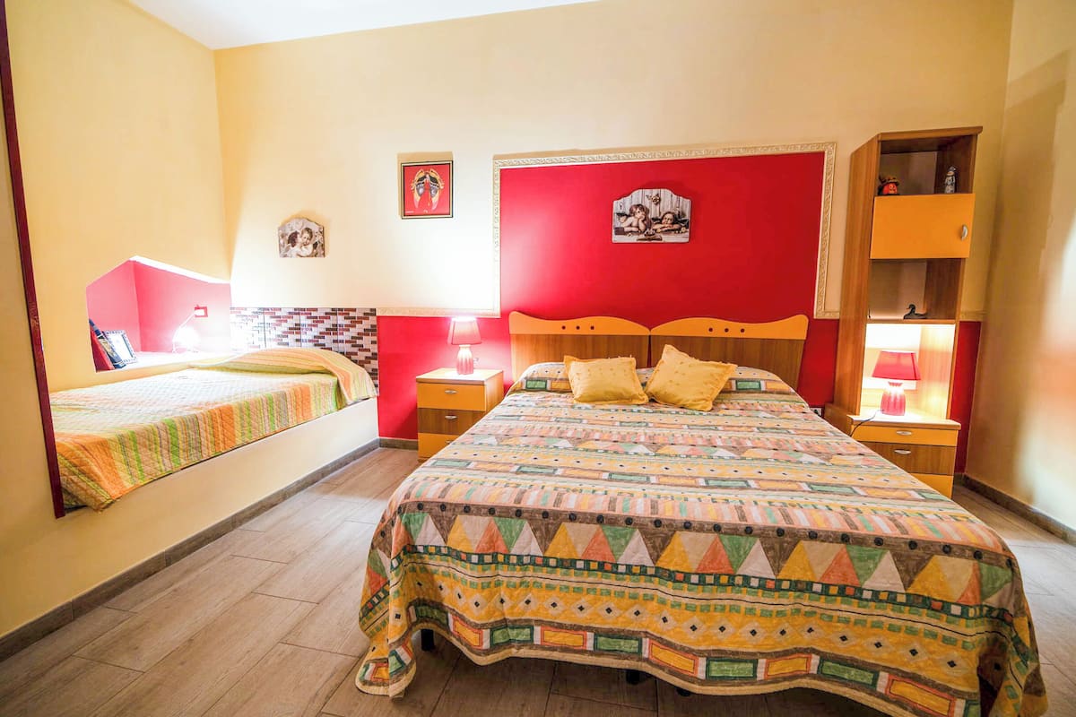 A bedroom with two beds, a red headboard, a bedside table with a lamp, and a small shelf with decorative items.