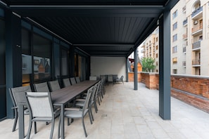 Un patio techado al aire libre con una mesa larga y sillas, una pared de ladrillo y vistas de un edificio residencial.