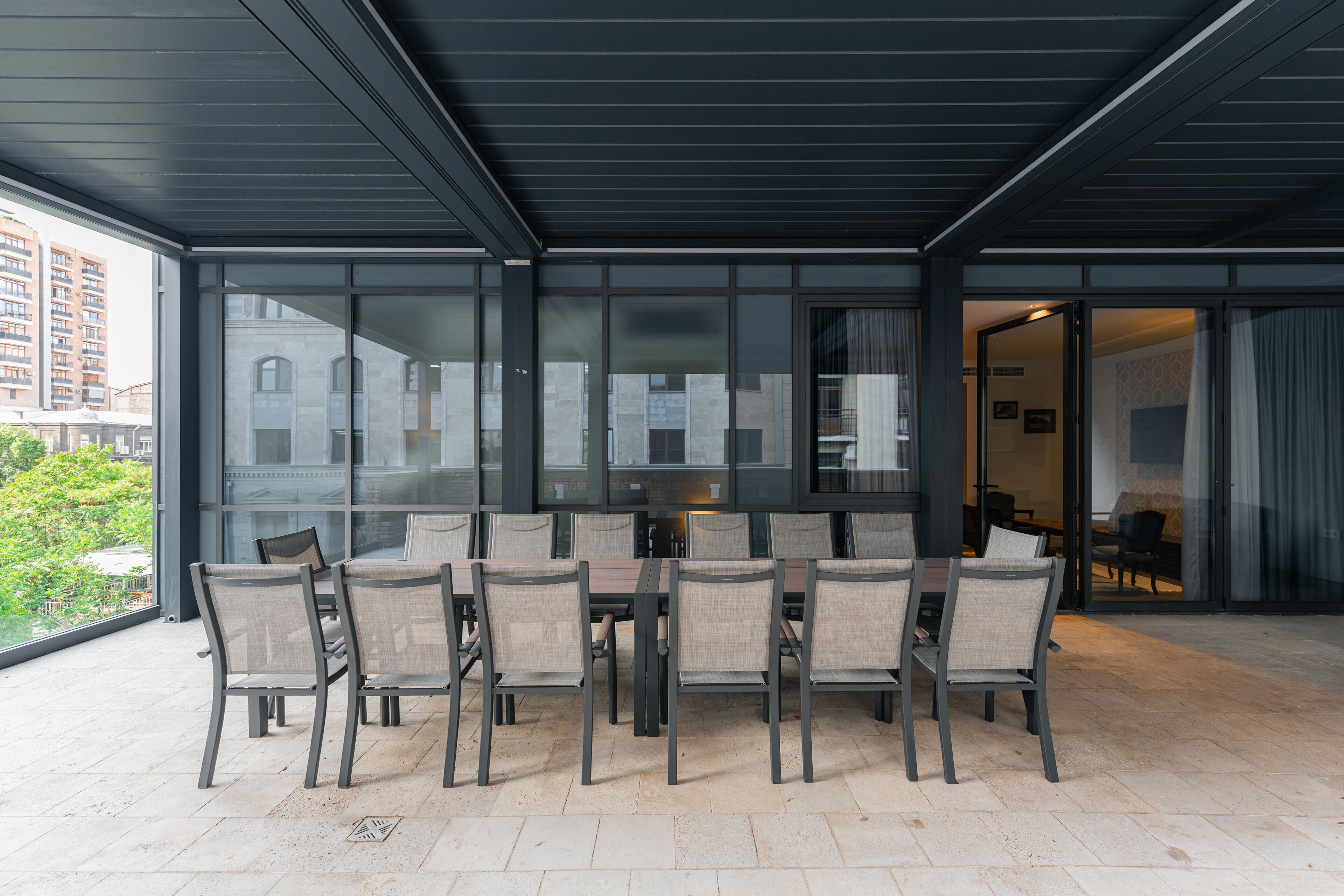 Un espace extérieur abrité, aménagé pour les repas, avec une table et des chaises, de grandes fenêtres et une vue sur les bâtiments.