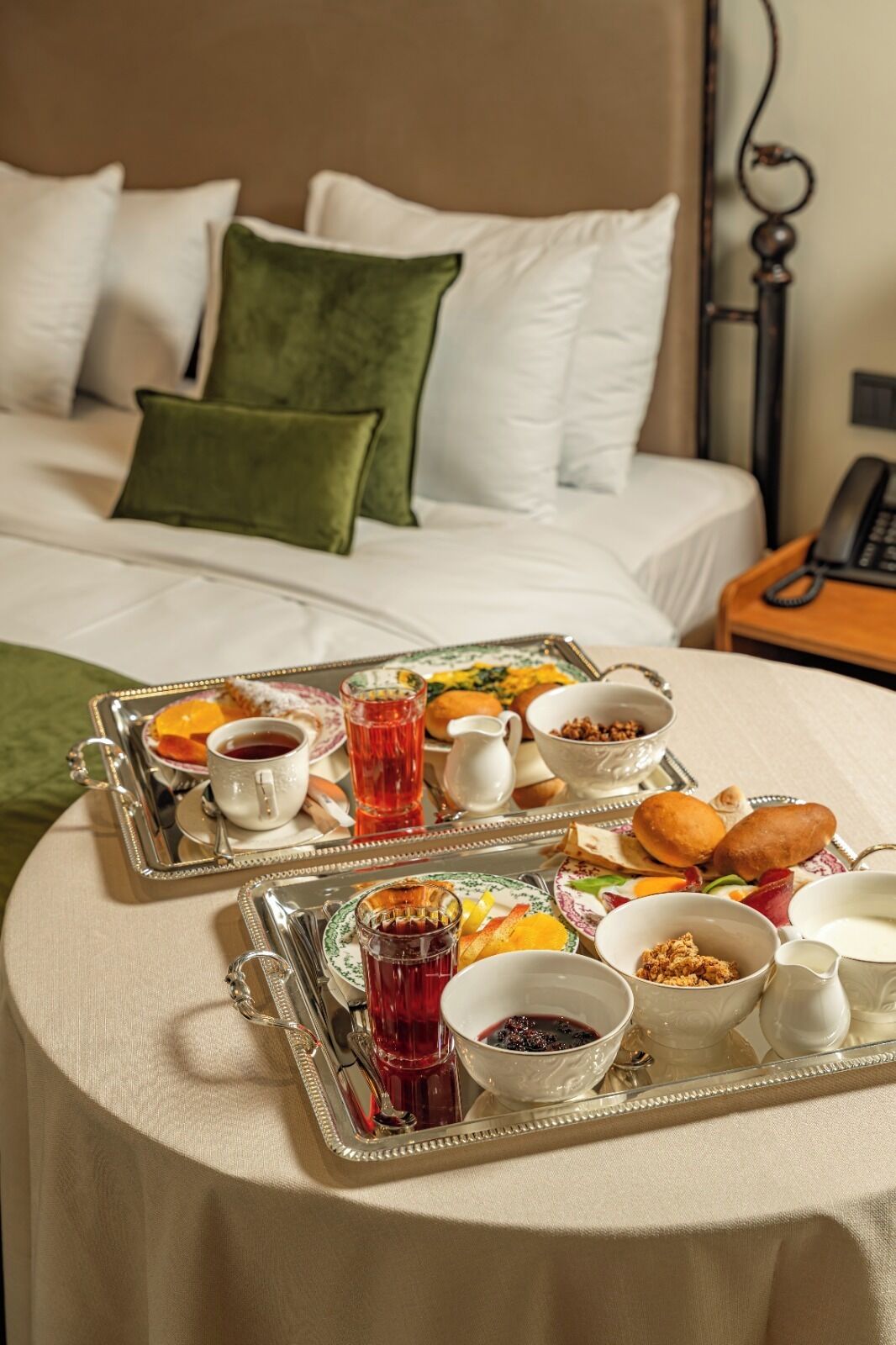 A neatly made bed with white and green pillows, a bedside table with a telephone, and a breakfast tray with various food items and drinks on a round table.
